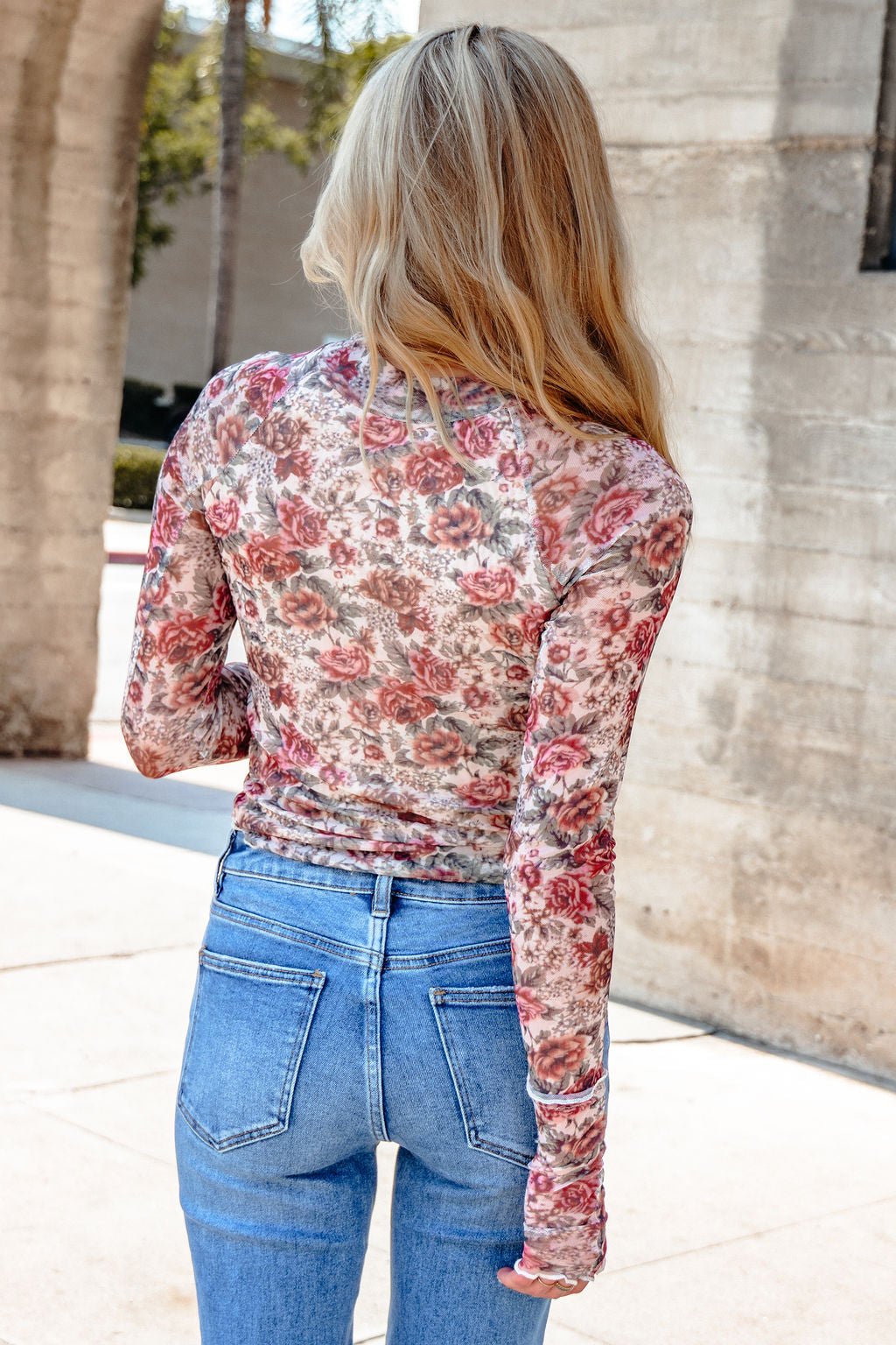 A woman with long blonde hair wears the Cream Floral Mesh Mock Neck Top and blue jeans, standing outdoors facing away.