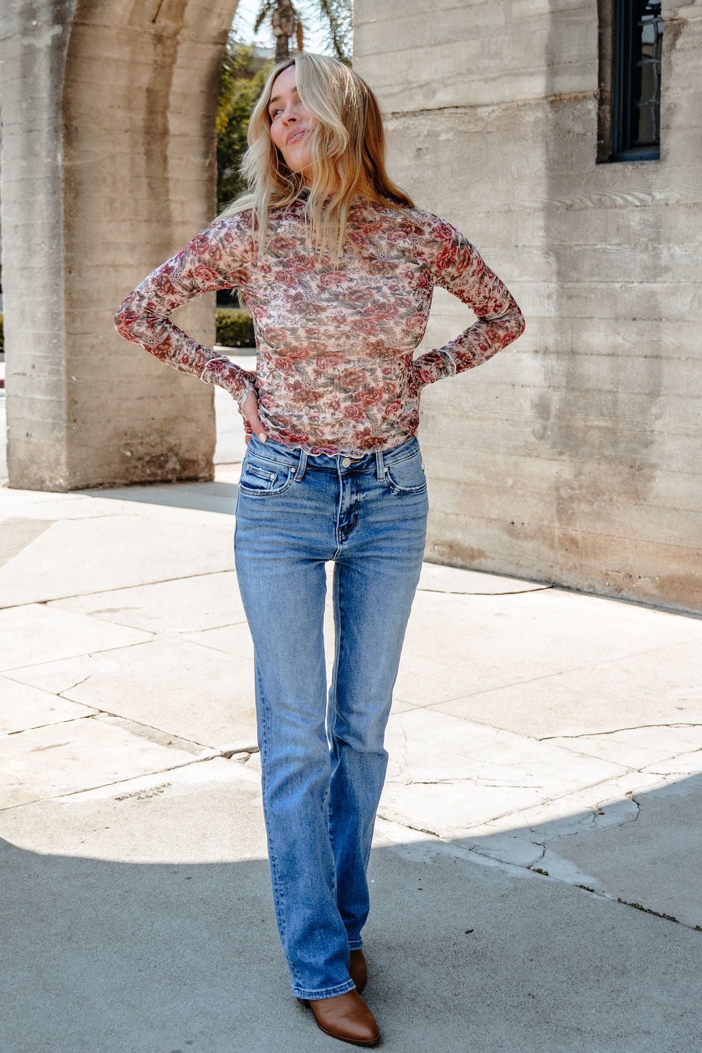 Woman outdoors in sunlight wearing the Cream Floral Mesh Mock Neck Top - DOORBUSTER, blue jeans, brown shoes, hands on hips.