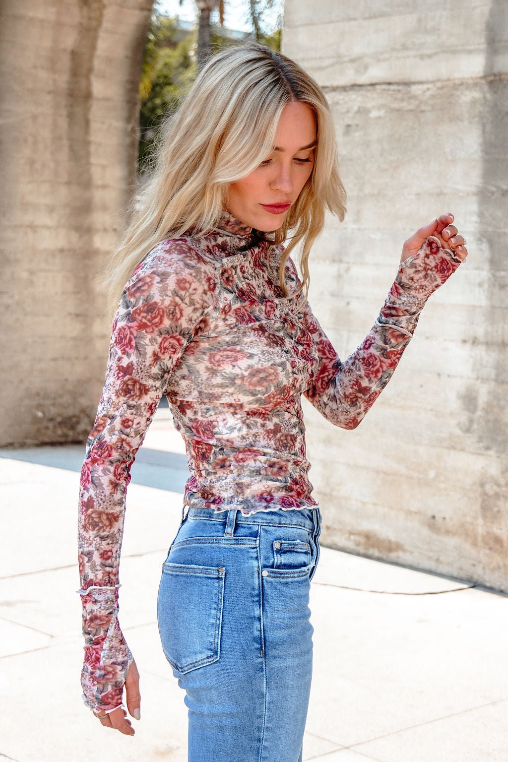A woman with long blonde hair wears the Cream Floral Mesh Mock Neck Top and blue jeans, standing outdoors near concrete structures.
