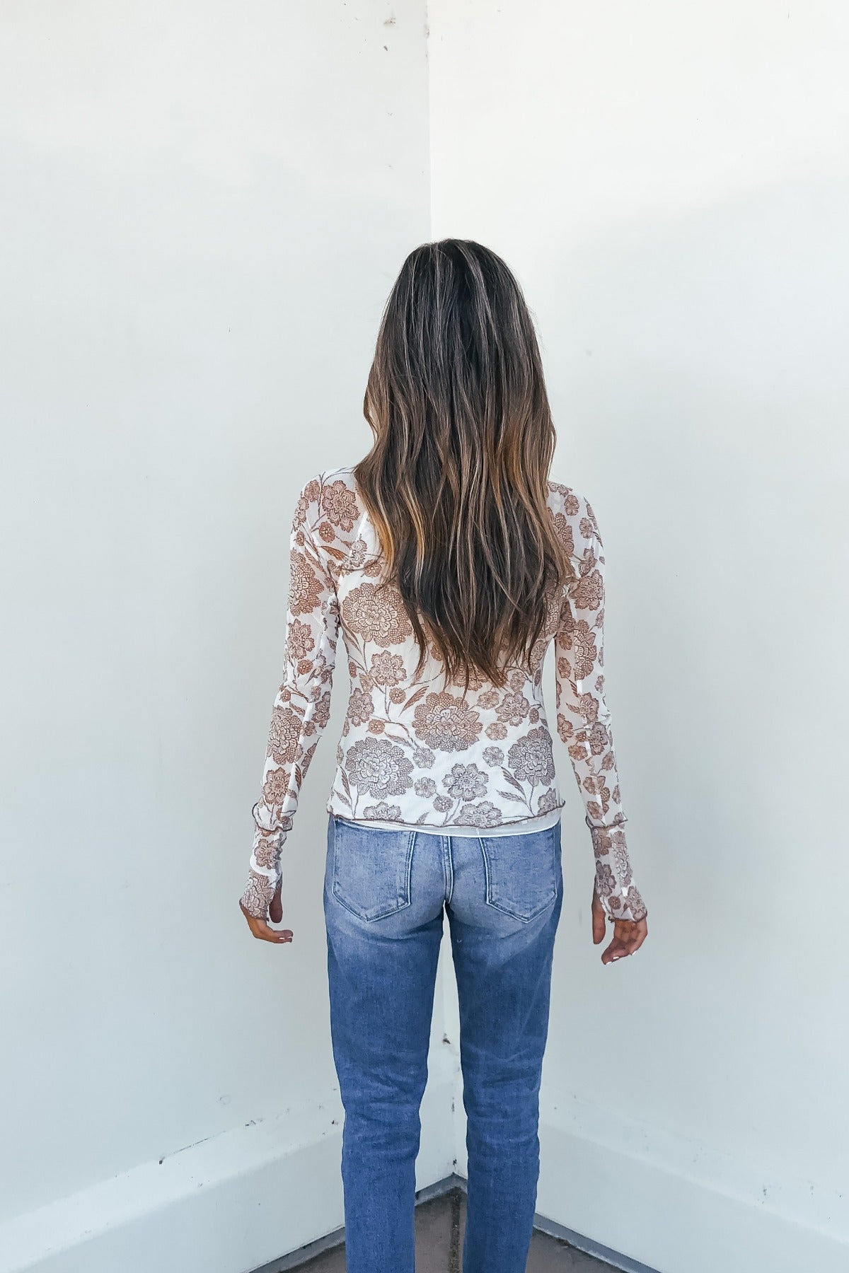 Woman with long wavy hair faces a white wall, wearing a Cream Floral Mesh Top and blue jeans.