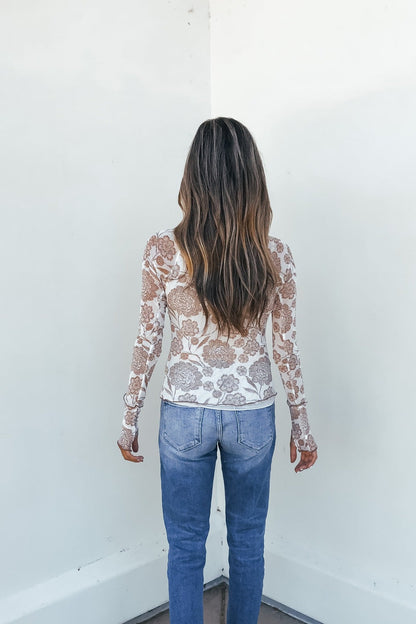 Woman with long wavy hair faces a white wall, wearing a Cream Floral Mesh Top and blue jeans.