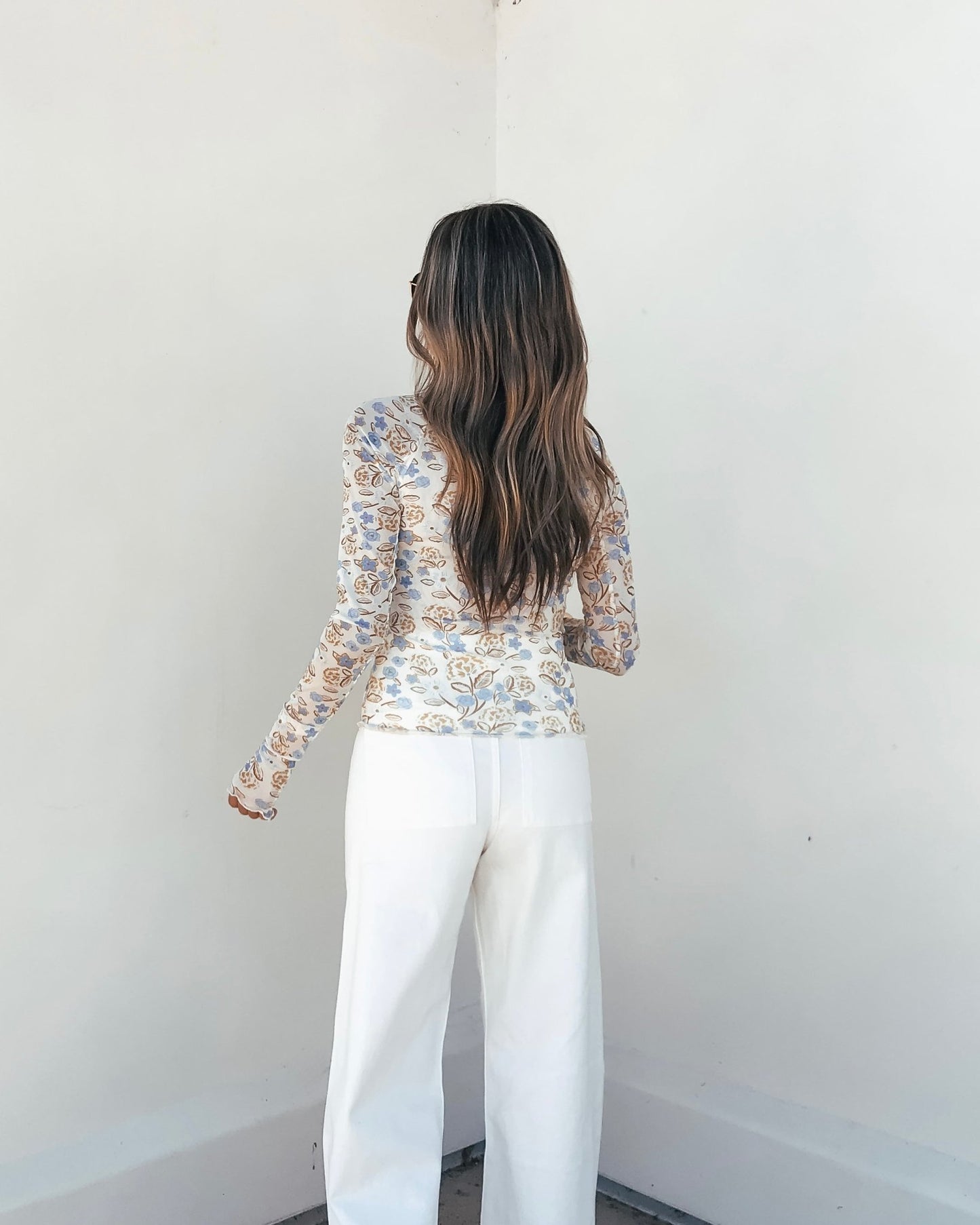 A woman with long wavy hair models the Cream Floral Mesh Top with sheer sleeves and white pants against a plain white wall.