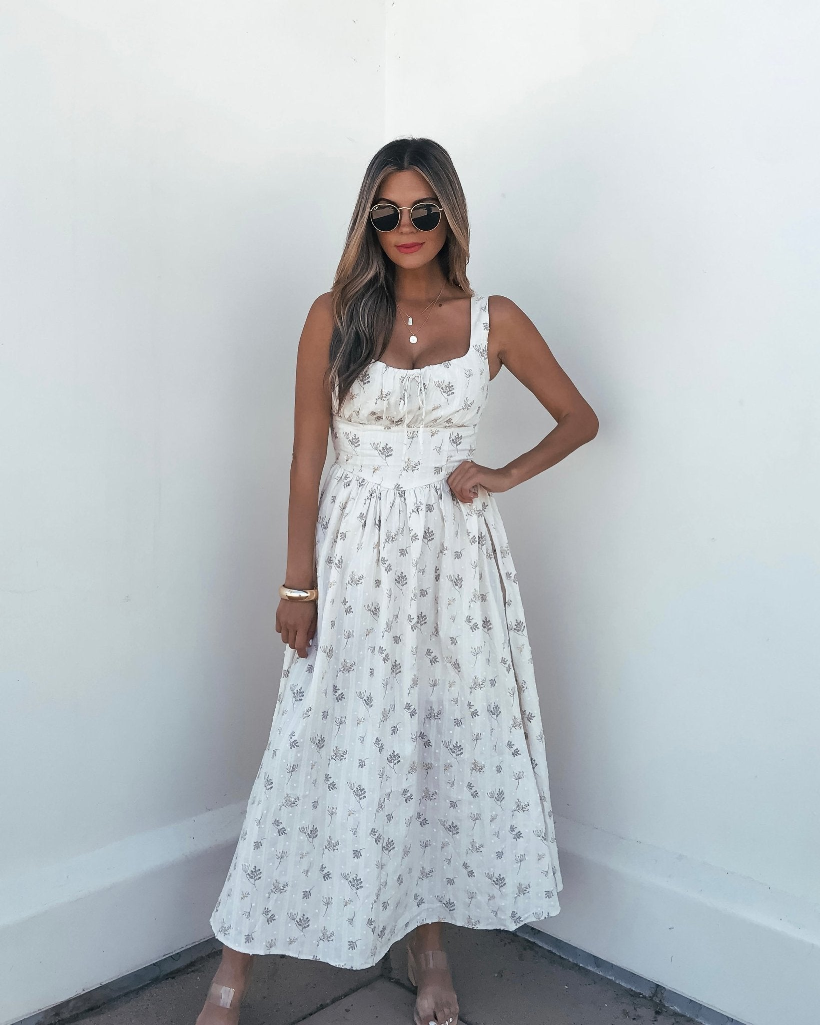 A woman wearing sunglasses and a Cream Floral Print Smocked Midi Dress poses with one hand on her waist against a white wall.