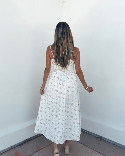 A woman wears the Cream Floral Print Smocked Midi Dress and sandals, facing a white wall with her back to the camera.