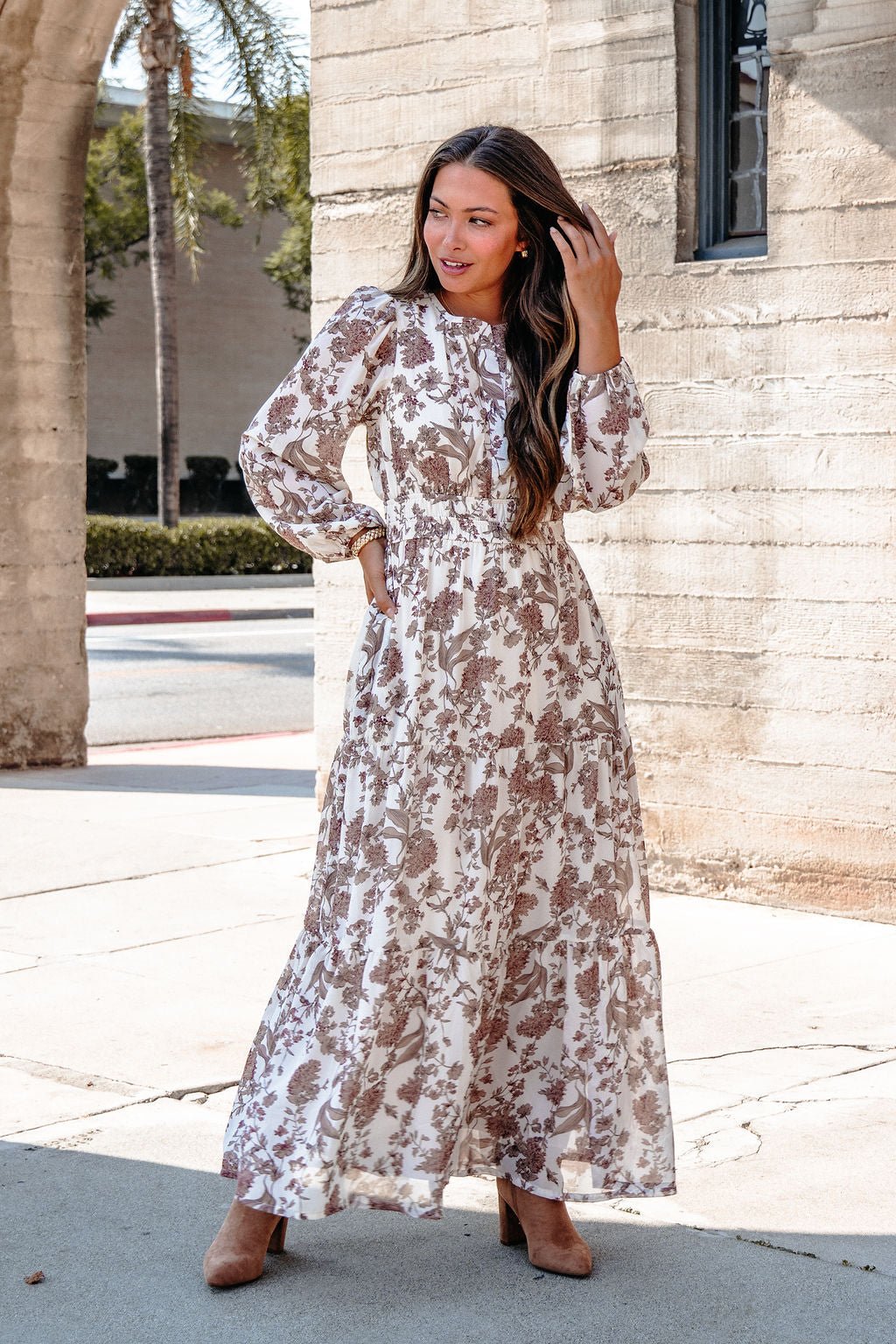 Woman in a Cream Floral Print Tiered Maxi Dress and brown heels stands by a stone building, touching her hair and smiling.