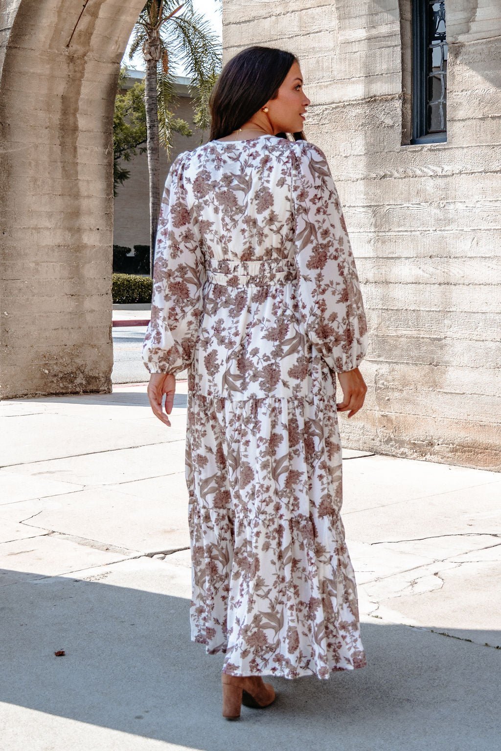 A woman in a Cream Floral Print Tiered Maxi Dress stands outdoors near stone arches on a sunny day, facing away.