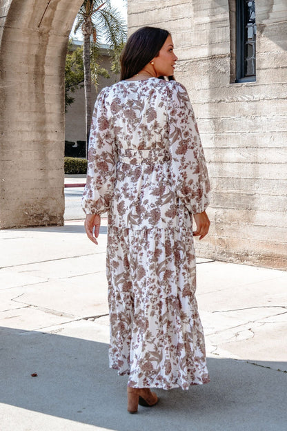 A woman in a Cream Floral Print Tiered Maxi Dress stands outdoors near stone arches on a sunny day, facing away.