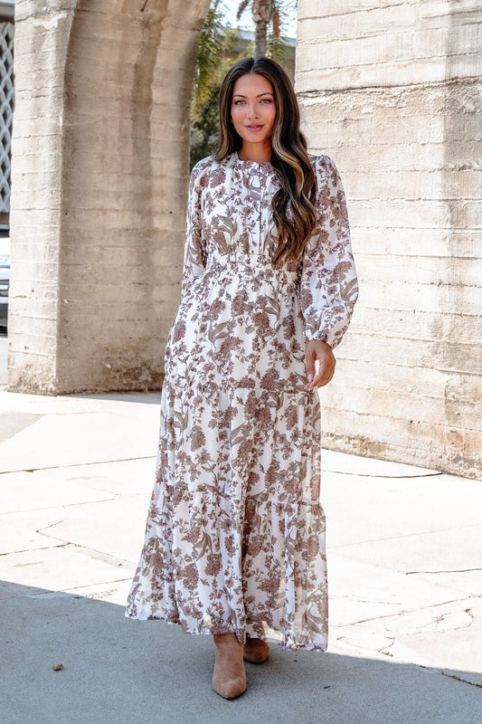 A woman in a Cream Floral Print Tiered Maxi Dress with brown heels stands outdoors by a concrete wall.