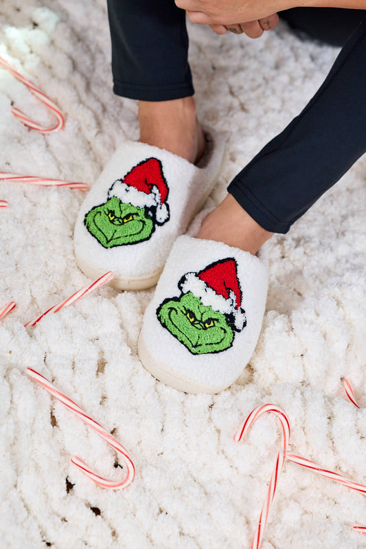 Person wearing Cream Grinch Plush Slippers with a Santa hat, standing on white carpet with candy canes—a great holiday gift.
