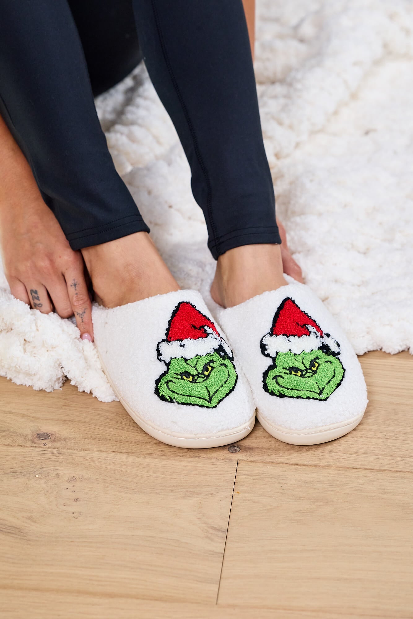 Person wearing Cream Grinch Plush Slippers - FINAL SALE sits on a fluffy rug—perfect holiday gift for any Grinch fan.