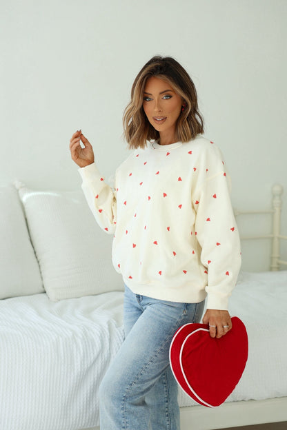 Woman in a Cream Heart Print Sweatshirt holding a red heart pillow, standing by a white bed.