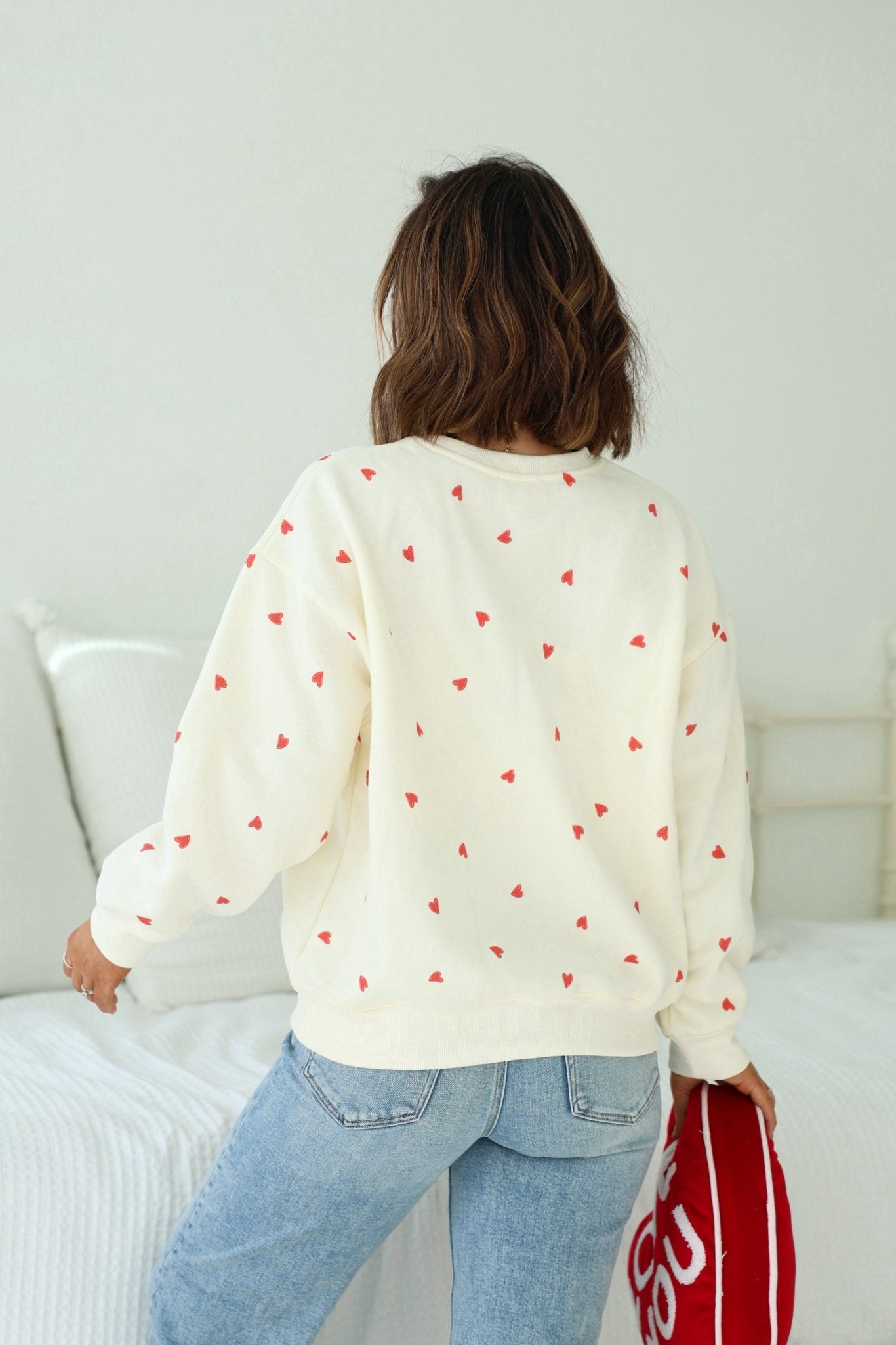 A person in a Cream Heart Print Sweatshirt and jeans stands by a white bed, holding a red bag.