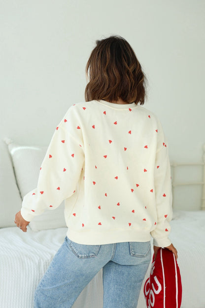 A person in a Cream Heart Print Sweatshirt and jeans stands by a white bed, holding a red bag.