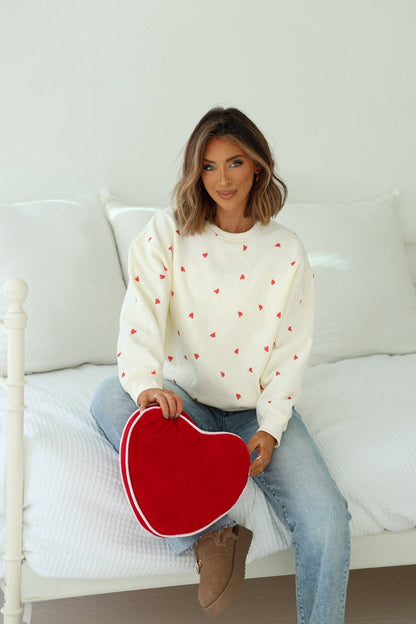 Woman on white bed, smiling at camera in a Cream Heart Print Sweatshirt, holding a red heart-shaped pillow.