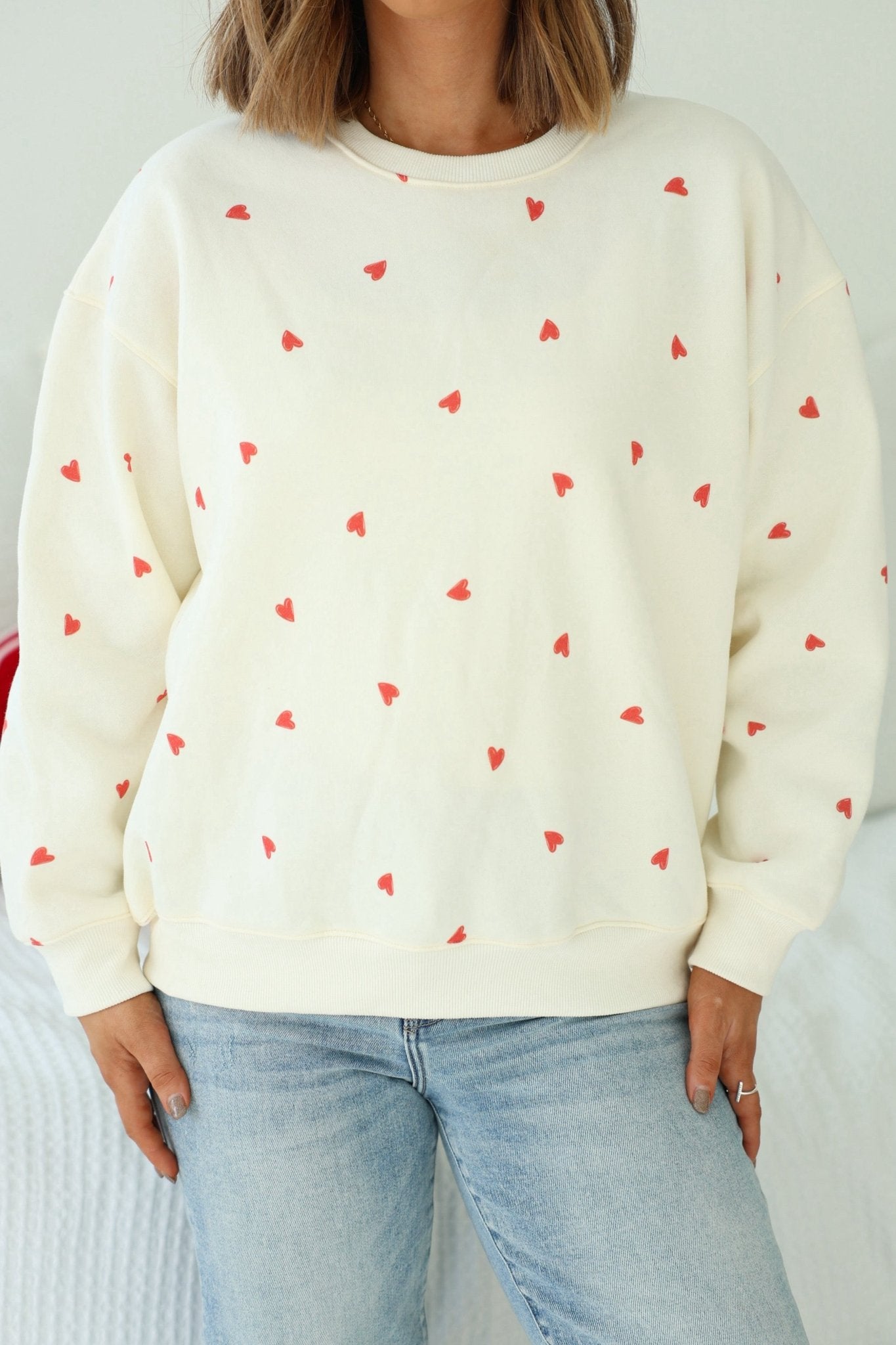 A person wearing a Cream Heart Print Sweatshirt with light blue jeans, standing indoors.