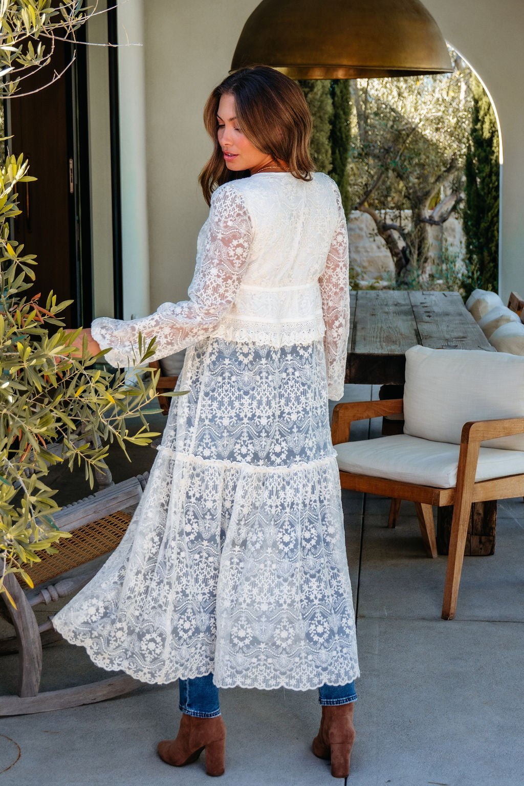 A woman wears a Cream Lace Tie Duster Cardigan over jeans and brown boots, standing outdoors and looking over her shoulder on a patio.