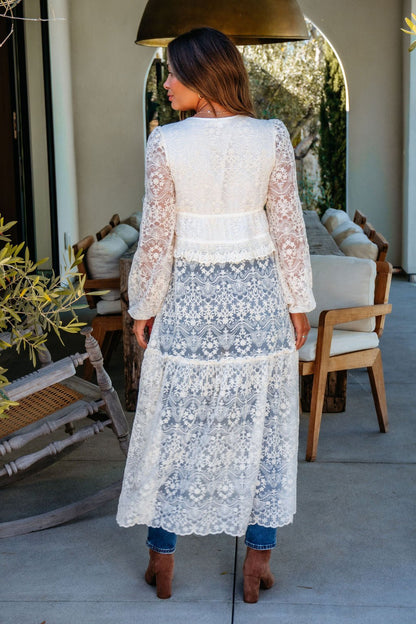 A woman wears the Cream Lace Tie Duster Cardigan over jeans and brown boots, standing outdoors with her back to the camera.