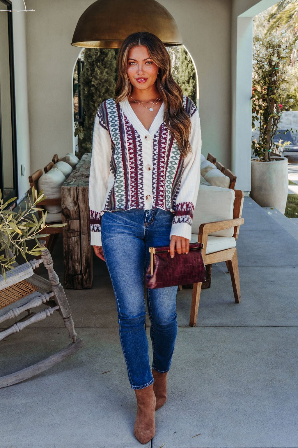 A woman outdoors in a Cream Multi Ethnic Print Button Down Cardigan holds a maroon clutch by patio furniture.