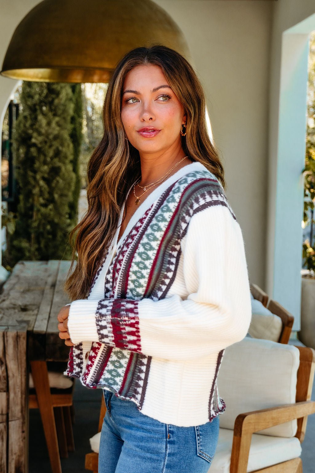 A woman with long, wavy hair models the Cream Multi Ethnic Print Button Down Cardigan, styled with jeans indoors by a wooden table.
