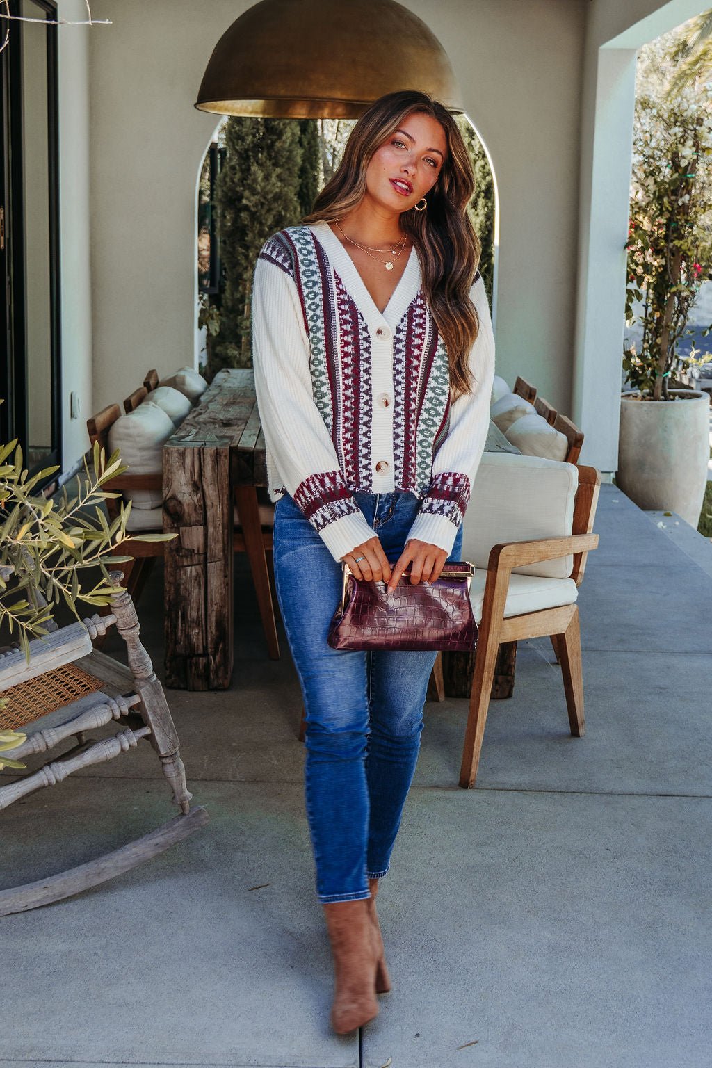 Woman stands on a patio in a Cream Multi Ethnic Print Button Down Cardigan, blue jeans, brown boots, and holding a clutch.