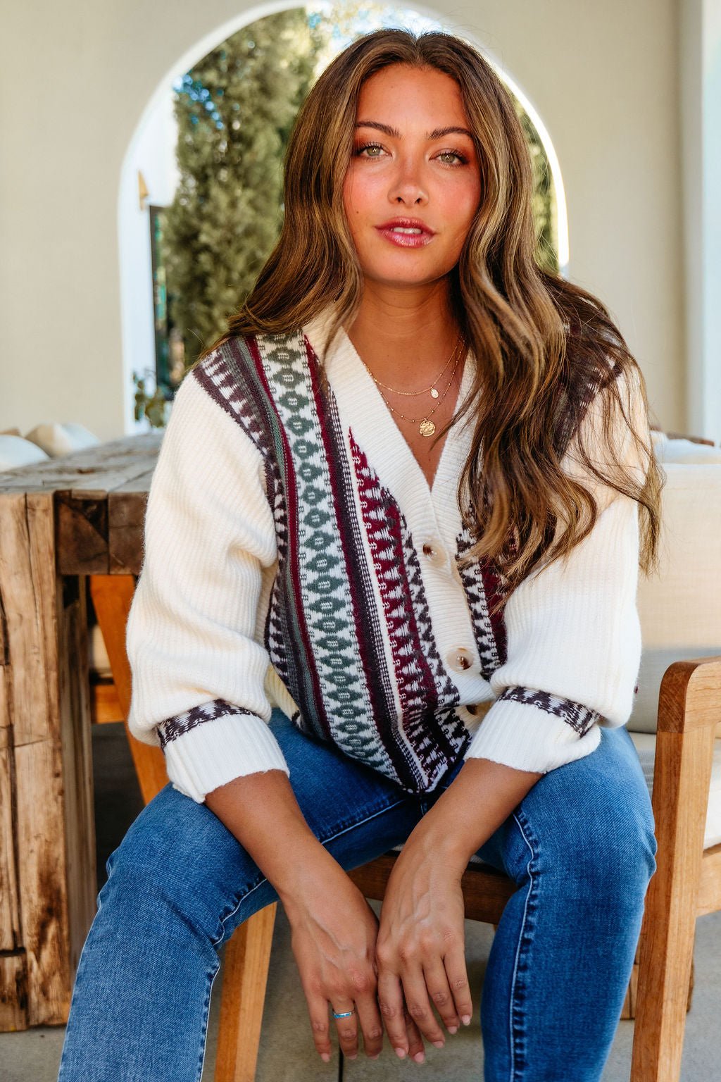 Woman with long brown hair wears the Cream Multi Ethnic Print Button Down Cardigan, seated outdoors and looking at the camera.