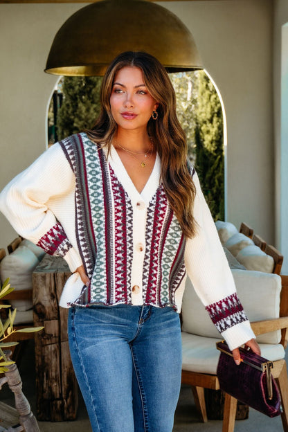 A woman wears the Cream Multi Ethnic Print Button Down Cardigan, blue jeans, and holds a clutch indoors, looking to the side.