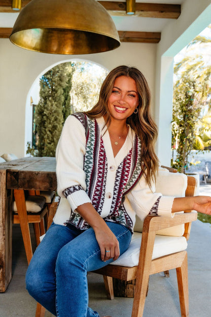 A smiling woman wears the Cream Multi Ethnic Print Button Down Cardigan, sitting outdoors at a wooden table in a bright space.