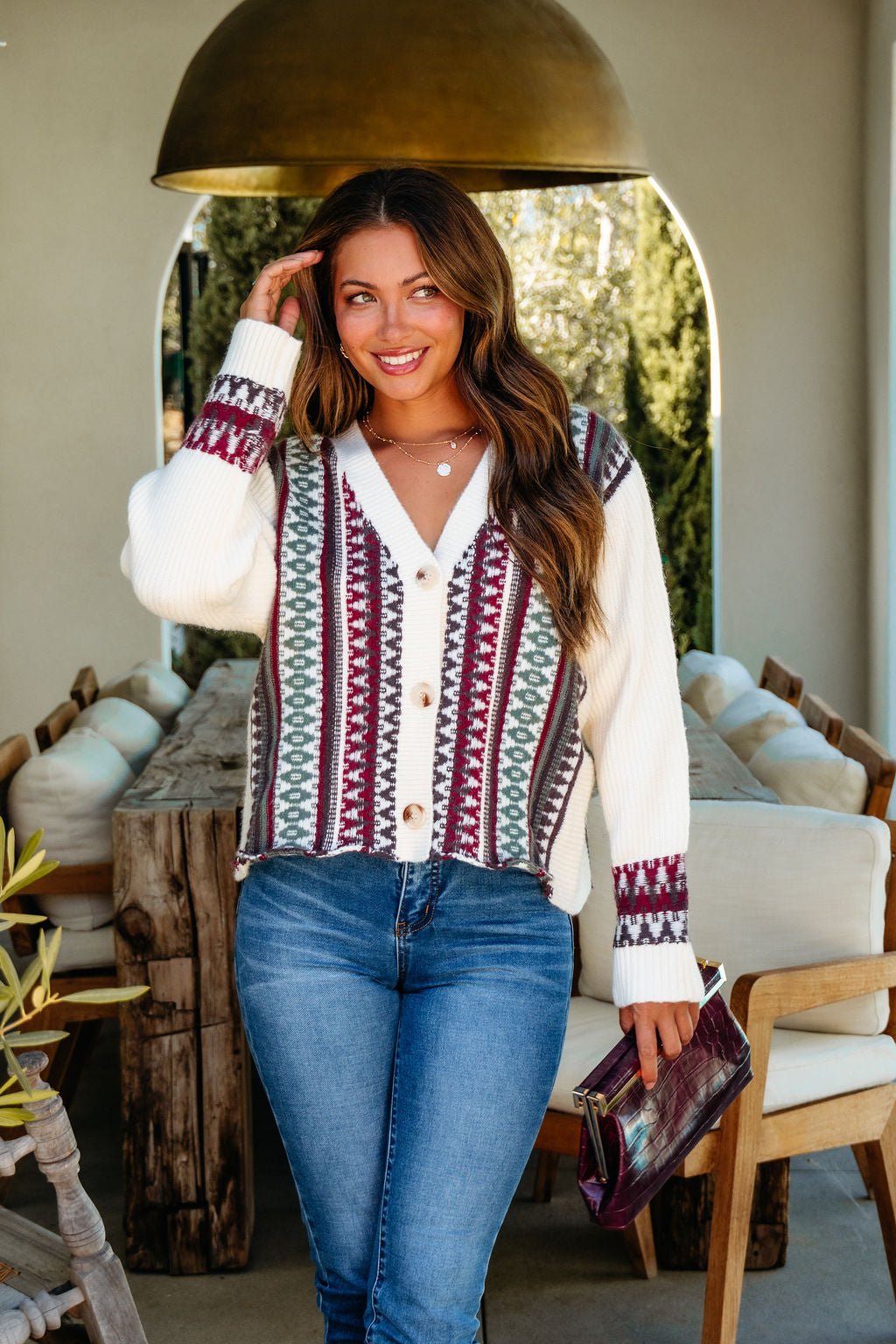 A smiling woman wears the Cream Multi Ethnic Print Button Down Cardigan indoors, paired with jeans, holding a purse.