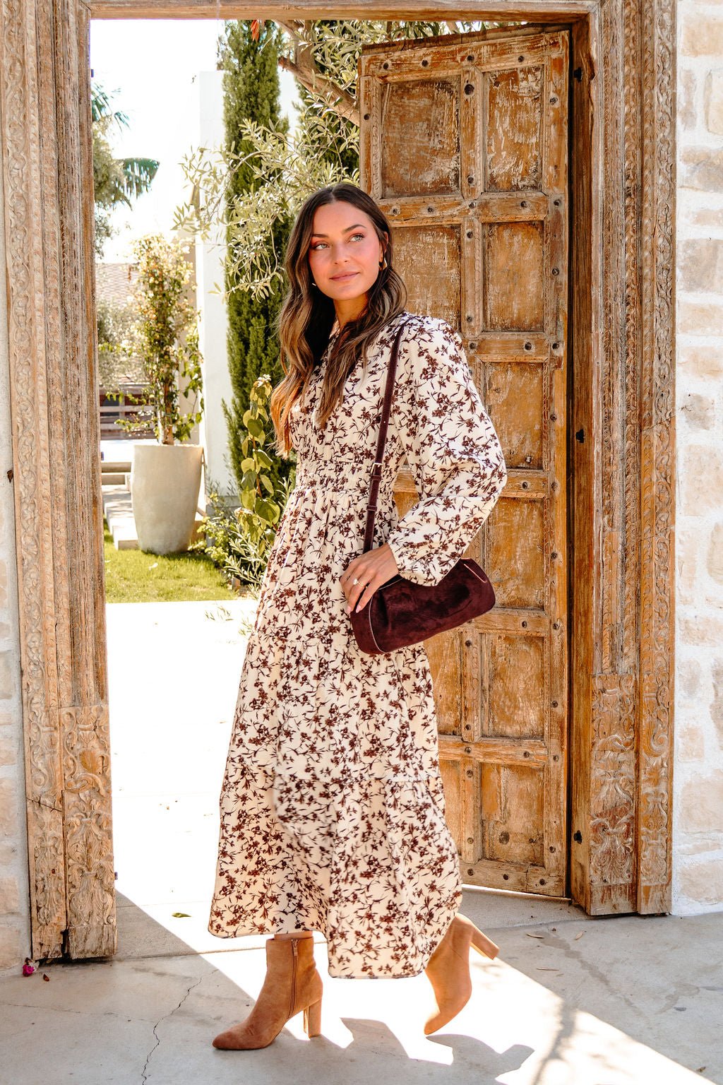 A woman in a Cream Multi Floral Print Tiered Midi Dress and boots stands in a rustic doorway with sunlight streaming in.