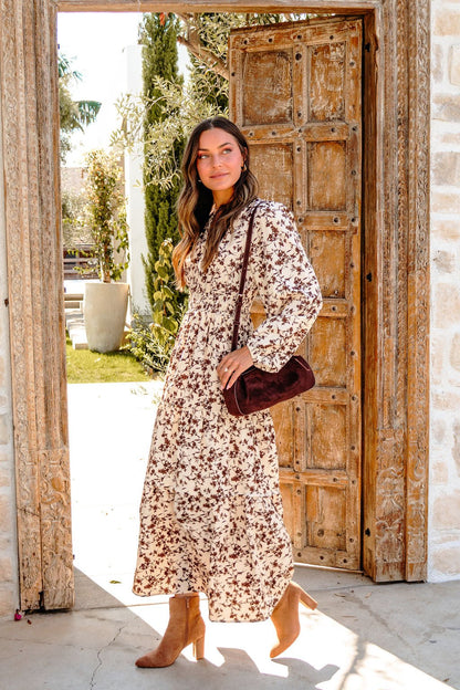 A woman in a Cream Multi Floral Print Tiered Midi Dress and boots stands in a rustic doorway with sunlight streaming in.