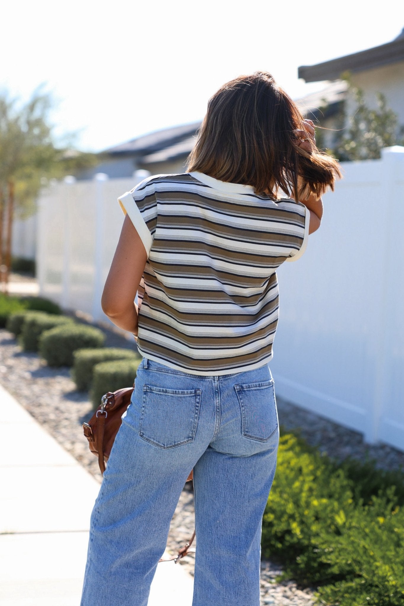 A woman with brown hair stands outside, facing away, wearing a Cream Multi Striped Cap Sleeve Top and jeans.