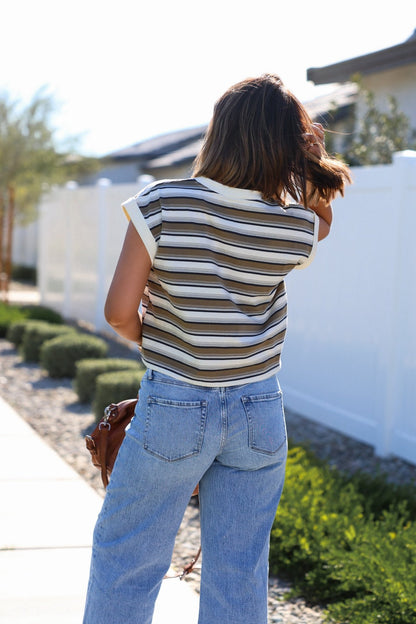 A woman with brown hair stands outside, facing away, wearing a Cream Multi Striped Cap Sleeve Top and jeans.