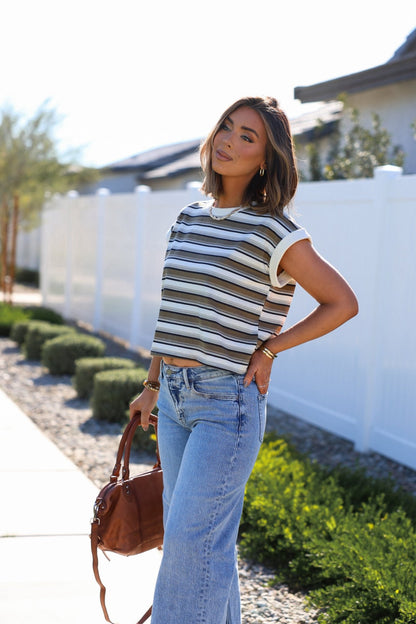 A woman wears a Cream Multi Striped Cap Sleeve Top and jeans, holding a brown handbag outdoors by a white fence and greenery.