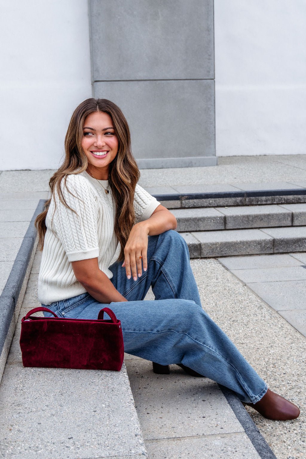 A woman in a Cream Puff Sleeve Cable Knit Sweater and jeans sits on outdoor steps, smiling, with a red handbag beside her.