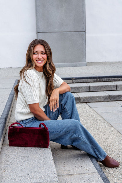 A woman in a Cream Puff Sleeve Cable Knit Sweater and jeans sits on outdoor steps, smiling, with a red handbag beside her.