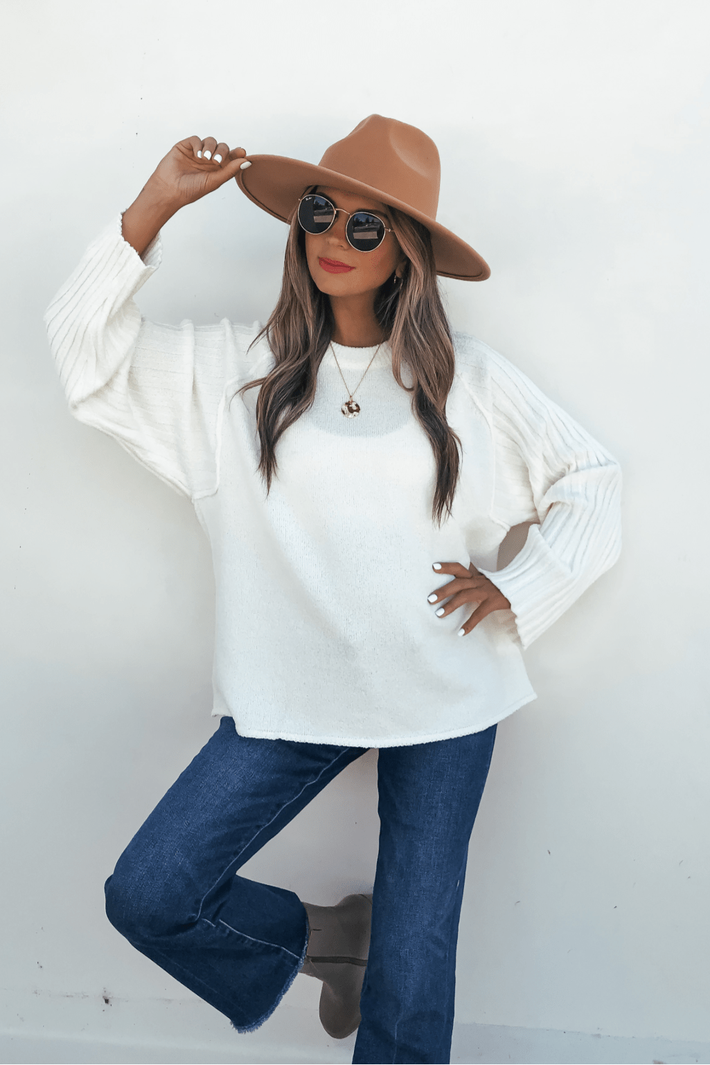 Woman poses against a white wall wearing sunglasses, tan hat, blue jeans, brown boots, and a Cream Raglan Sleeve Textured Sweater.