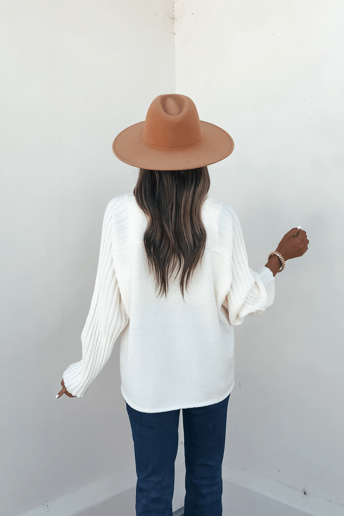 A woman faces away in a cream raglan sleeve textured sweater, brown hat, and dark jeans against a plain light-colored wall.