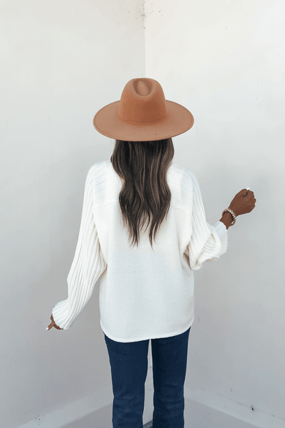A woman faces away in a cream raglan sleeve textured sweater, brown hat, and dark jeans against a plain light-colored wall.