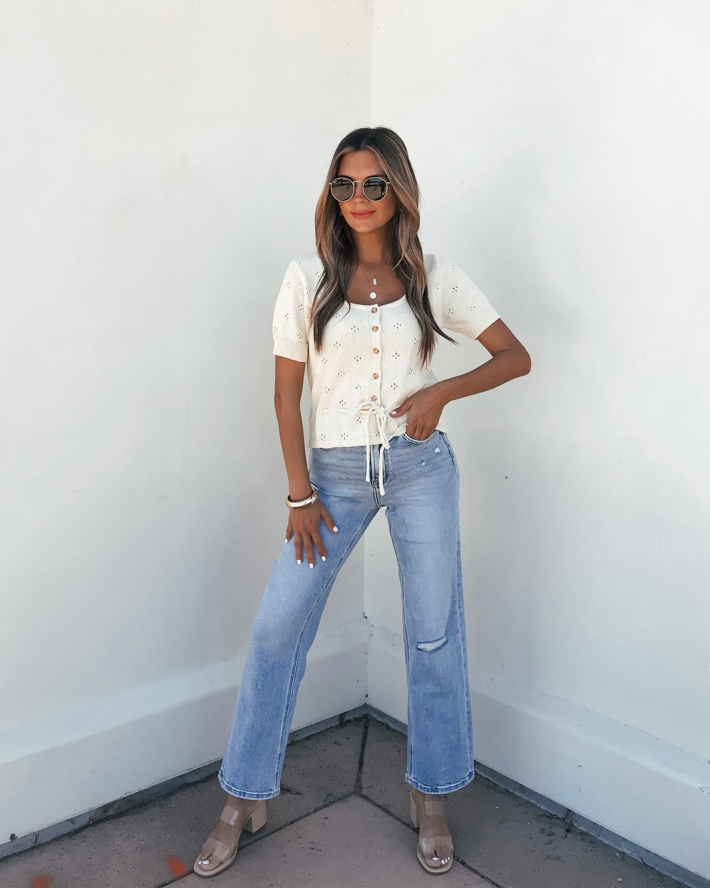 Woman in sunglasses, a white short-sleeve blouse, light blue jeans, Cream Tie Button Up Sweater, and clear heels poses confidently.