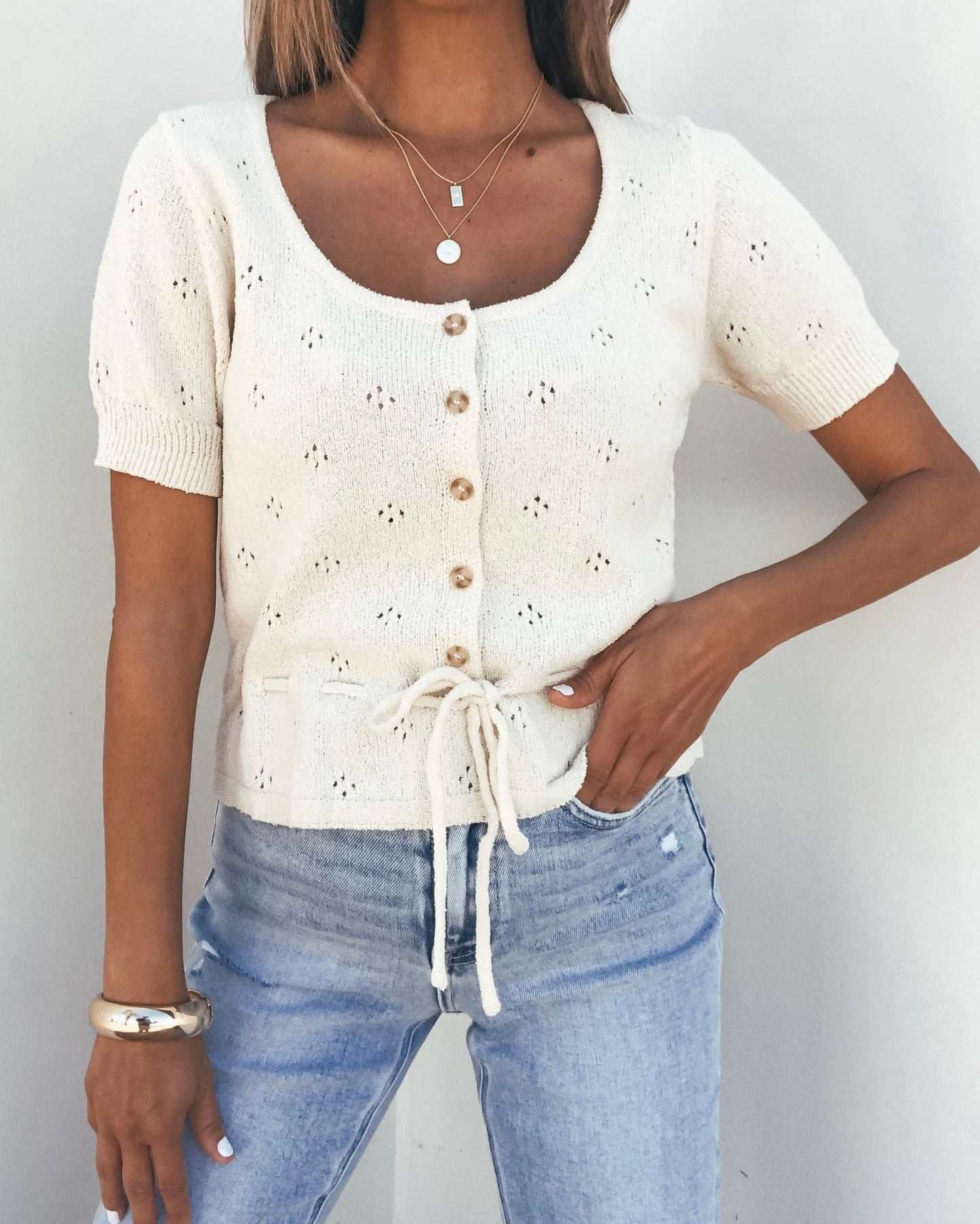 Woman wearing a Cream Tie Button Up Sweater, light blue jeans, layered necklaces, and bracelets.
