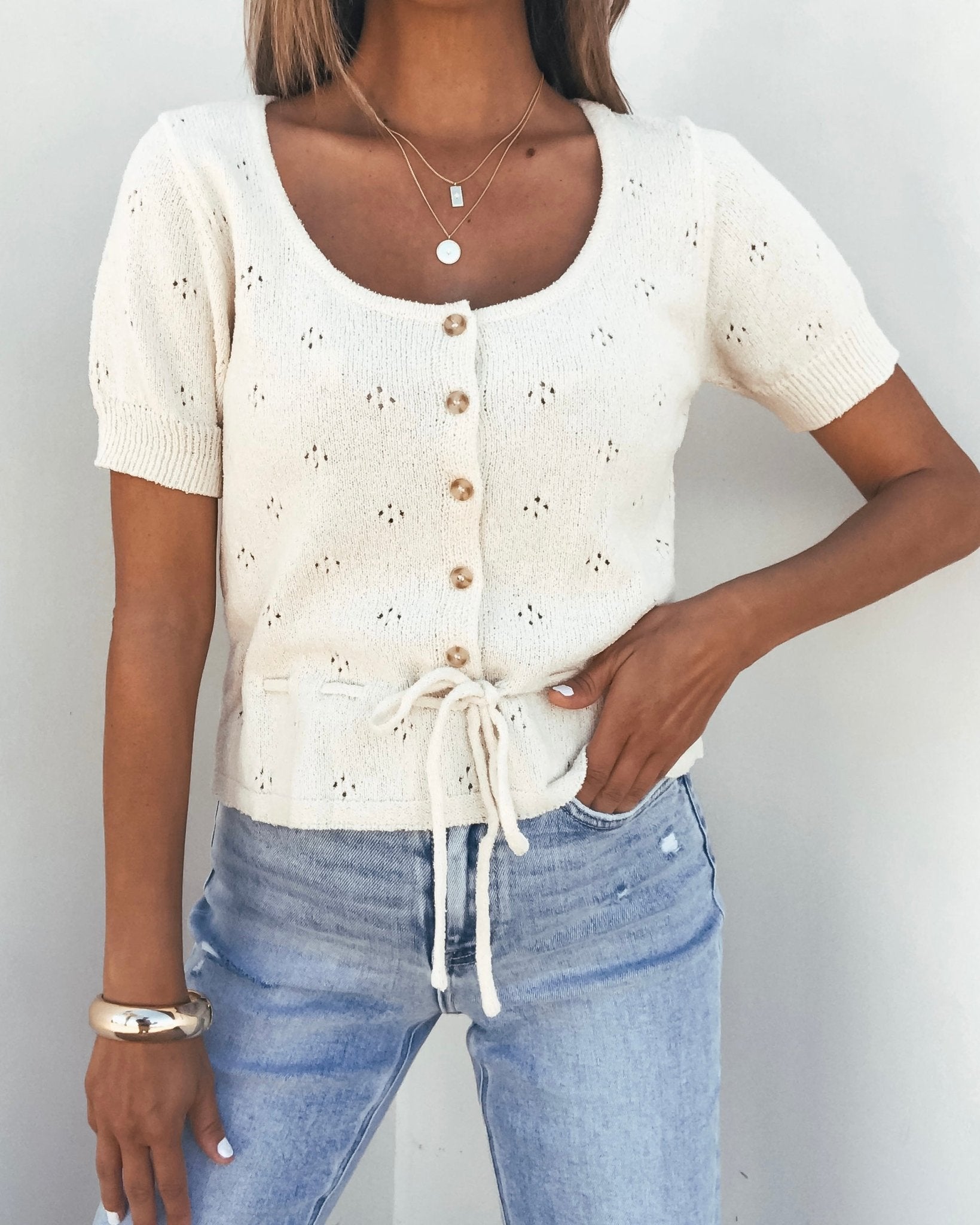 Woman wearing a Cream Tie Button Up Sweater, light blue jeans, layered necklaces, and bracelets.
