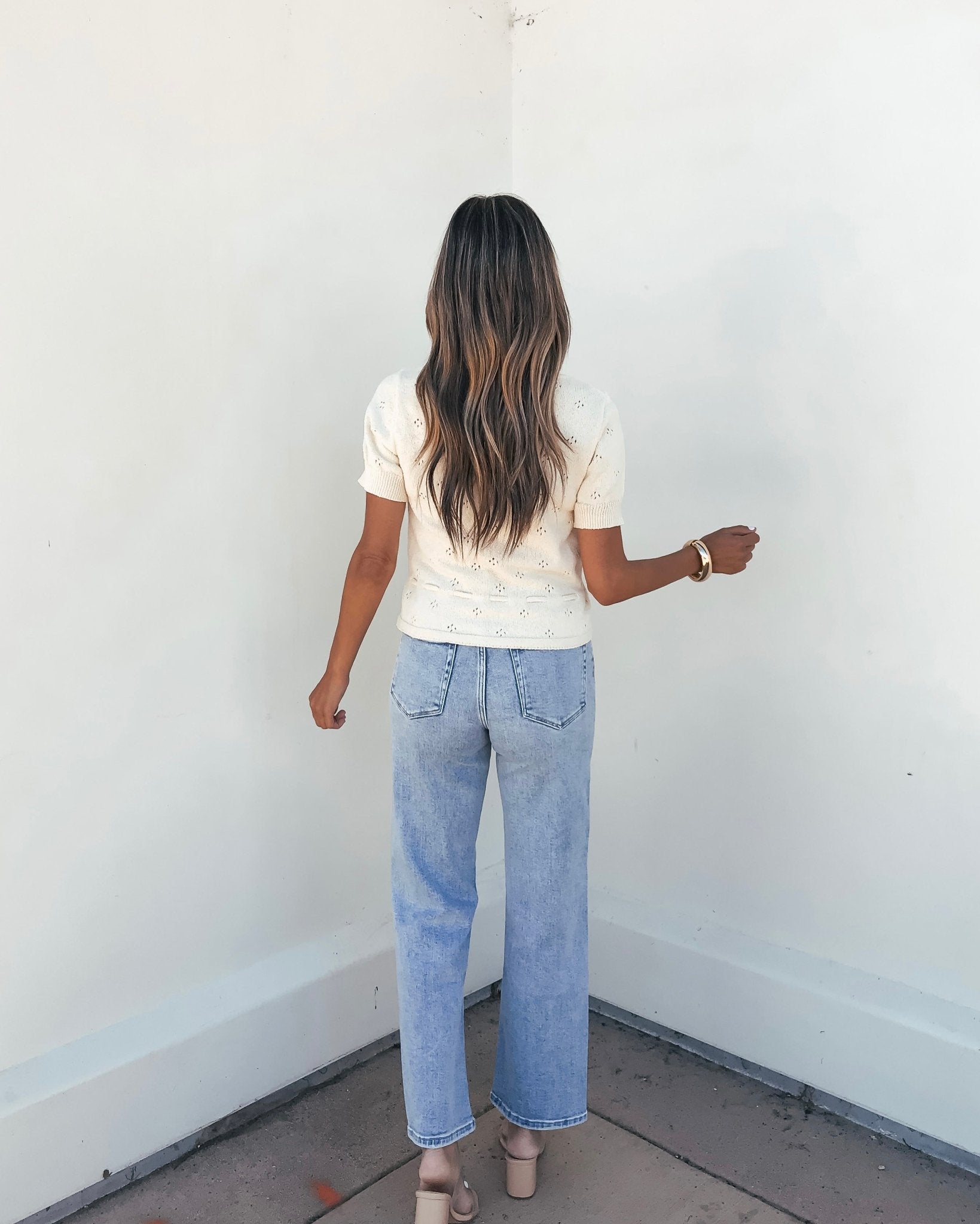 A woman with long wavy hair faces a white wall, wearing the Cream Tie Button Up Sweater, light blue jeans, and beige heels.