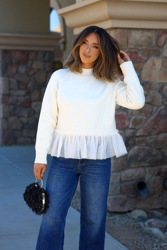 Woman in a Cream Tulle Mesh Peplum Sweater - FINAL SALE, blue jeans, and black purse, standing by a stone wall.