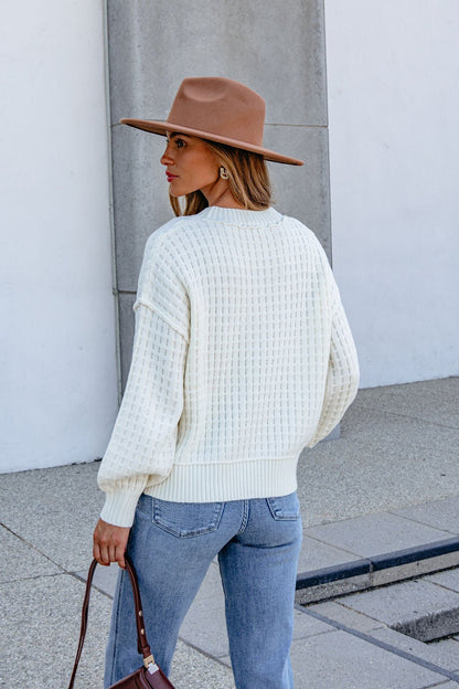 Woman in a Cream Waffle Knit Sweater, blue jeans, and brown hat stands outside with a brown handbag, facing away.