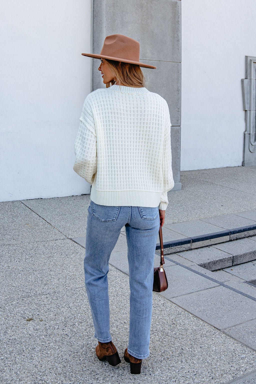 A woman in a Cream Waffle Knit Sweater, jeans, and brown hat holds a brown purse while standing on a sidewalk, facing away.