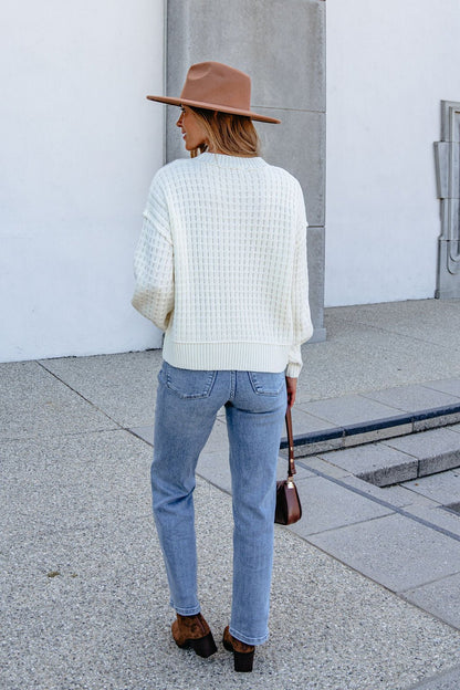 A woman in a Cream Waffle Knit Sweater, jeans, and brown hat holds a brown purse while standing on a sidewalk, facing away.