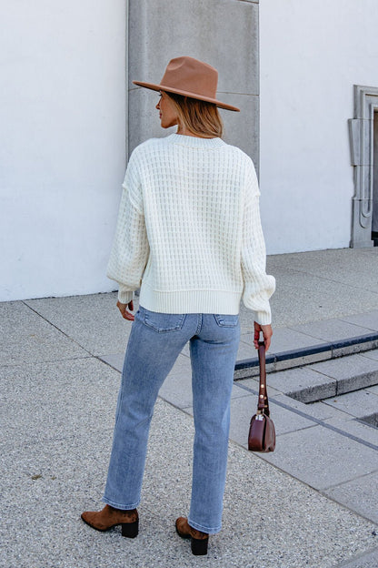 Woman in Cream Waffle Knit Sweater, blue jeans, brown hat, and boots stands outside with a small brown purse, facing away.