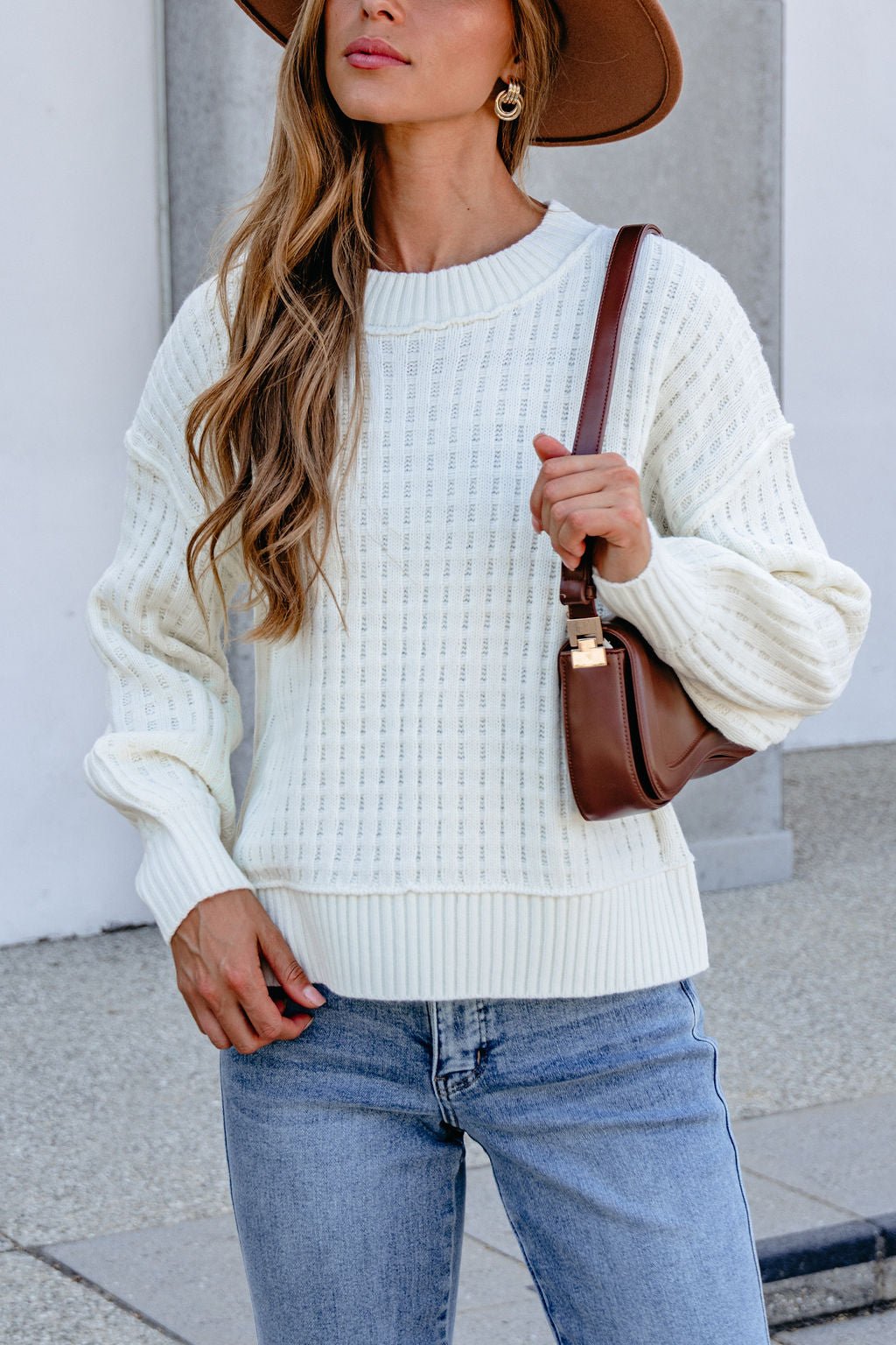 Woman in a Cream Waffle Knit Sweater, light blue jeans, and brown hat holds a brown shoulder bag while standing outdoors.