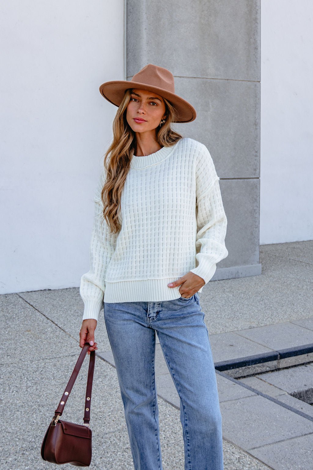 Woman in Cream Waffle Knit Sweater, blue jeans, and brown hat stands outdoors holding a brown handbag against a neutral background.