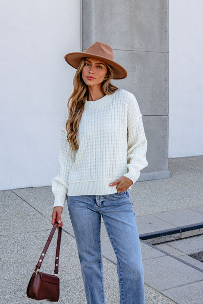 Woman in Cream Waffle Knit Sweater, blue jeans, and brown hat stands outdoors holding a brown handbag against a neutral background.